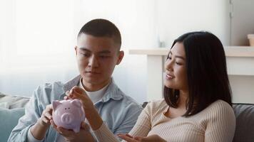 A young couple sits on a sofa, discussing their finances. One partner is holding a pink piggy bank, while the other gestures, suggesting plans and strategies for saving money. video