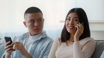 A woman smiles and talks on her phone while a man beside her looks at his device. Sunlight filters through sheer curtains, creating a warm atmosphere in the room. video