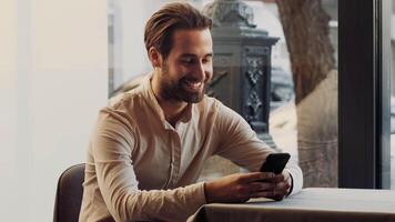 A man sits at a table in a bright cafe, smiling as he interacts with his smartphone. The warm sunlight streams through the windows, creating a welcoming atmosphere. video
