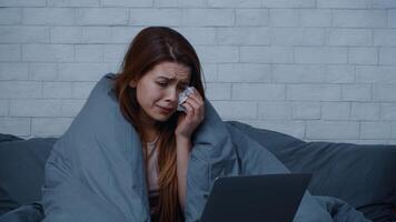 A woman wrapped in a blanket sits on a sofa, showing signs of distress as she wipes her tears. It is winter, and the atmosphere conveys feelings of loneliness and anxiety in her home setting. video