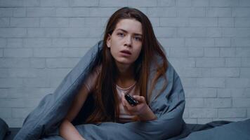 A Caucasian woman sits in bed wrapped in a blanket, staring blankly as she holds a remote control. This depicts feelings of loneliness and depression during winter at home. video