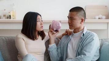 A couple sits on a couch in their living room, appearing to discuss finances. One person holds a pink piggy bank while looking at the other with a thoughtful expression. video