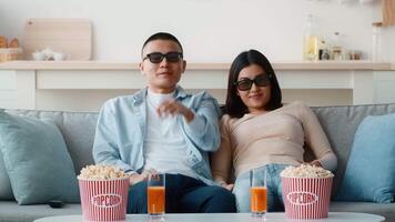 A couple sits together on a comfortable couch, wearing sunglasses, surrounded by bowls of popcorn and colorful drinks. They are ready for an entertaining movie night in their stylish living room. video