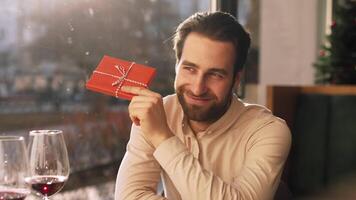 A cheerful man sits at a cafe table, holding a neatly wrapped gift box. The warm atmosphere and holiday decorations create a festive mood, enhancing his joyful expression. video