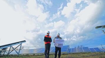 Engineers with laptop and project plan on the solar farm. Workers in special uniform standing among the solar panels and discuss the installation of sunny batteries. video