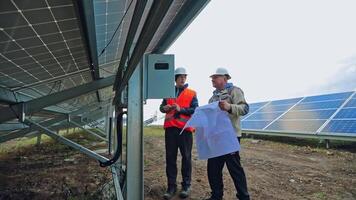 Solar technicians install photovoltaic panel on the field. Two engineers in protective uniform and hard hat with project plan under the big solar panel. video