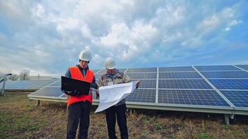 solar paneles instalación. dos trabajadores en el antecedentes de azul fotovoltaica panel. ingenieros en difícil sombrero mirando dentro el ordenador portátil y esquema plan en un solar granja. video