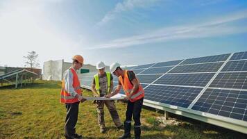 Male technicians with a project plan on a solar farm. Three workers in protective uniform and helmets discussing about solar panels installation in a bright day. video