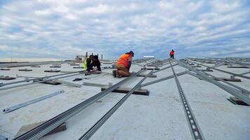 Installation of innovative solar farm under the blue sky. Metal basis for solar panels on roof. Workers in uniform construct base for photovoltaic cells. video