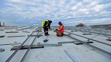 New construction of solar farm on the top of building. Two workers in colorful waistcoats attaching metal base on a flat roof. video