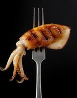 grilled squid on a sleek two-prong fork with charred grill marks and glossy texture for seafood presentations on dark background photo