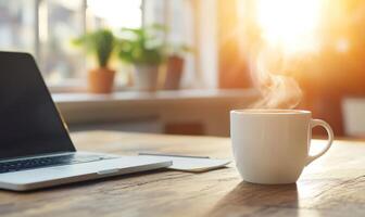 A steaming cup of coffee sits on a wooden desk next to a laptop and a notepad. Sunlight shines through the window behind the objects. photo