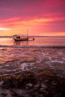 A fisherman's boat rests on the shore at Pasir Panjang Beach during sunset. The sky is adorned with clouds in beautiful red and yellow gradients, creating a stunning and tranquil scene photo
