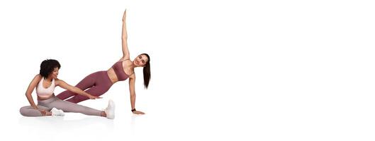 Two women engage in a fitness routine, demonstrating flexibility and strength. One performs a side plank while the other stretches her leg, both wearing athletic gear in a bright setting. photo