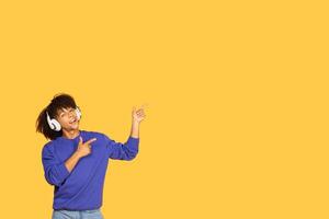 A joyful young man with headphones smiles while pointing confidently at copy space. Dressed casually in a blue sweater and jeans, he exudes a lively vibe against a vibrant yellow background. photo