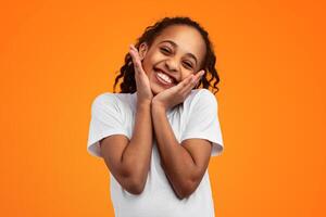 Portrait Of Cute Joyful African American Girl Touching Cheeks And Smiling At Camera, Young Child Posing Isolated Over Yellow Studio Background With Copy Space, Banner, Closeup photo