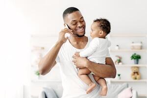 Multitasking Concept. Young Black Man Holding Little Baby And Talking On Cellphone While Working Remotely With Laptop At Home, African American Freelancer Guy Enjoying Paternity Leave, Copy Space photo