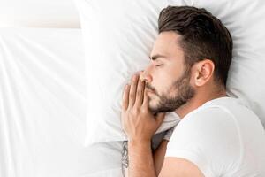 Asleep millennial man sleeping, resting peacefully in comfortable bed, lying with closed eyes, top view, free space. Recreation, deep male sleep, time to rest and nap concept photo