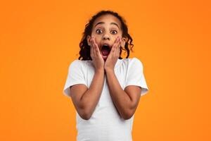 Omg. Portrait of excited surprised black girl touching her face and cheeks, looking at camera with open mouth, shocked by great offer, standing isolated over yellow orange studio background photo