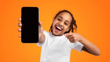 Trendy Mobile Phone. Excited young black girl holding smartphone with empty screen in hand, showing device close to camera and pointing at gadget with blank space, mockup, yellow studio background photo