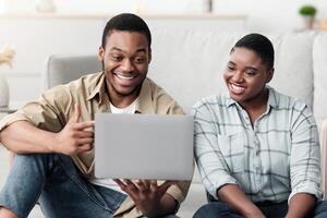 African American Family Couple Using Laptop Planning Vacation And Booking Tickets Online Sitting On Couch At Home, Pointing Finger At Computer. Black Spouses Choosing Travel Destination For Trip photo