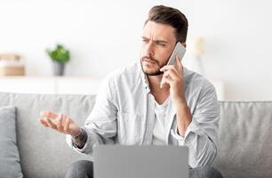 Unpleasant call. Worried freelancer having problems with internet connection while working on laptop, calling customer support on smartphone, sitting on sofa, free space photo