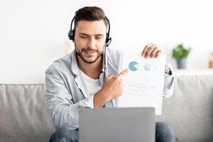 Online meeting. Happy young manager having call on laptop computer, showing diagram and market reports to webcam. Man wearing headset and looking at pc screen photo