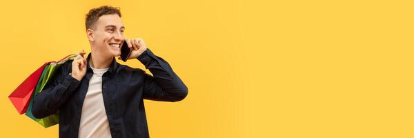 Positive handsome young guy wearing casual clothing with colorful shopping bags on his shoulder talking on phone and looking at copy space for advertisement, yellow background, web-banner photo
