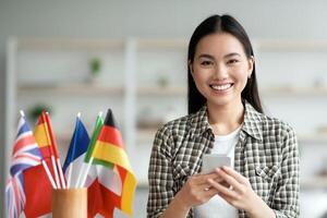 Cheerful asian young lady using smartphone, having English or other foreign language class, sitting at table with wireless headset and international flags at home, using mobile application, copy space photo