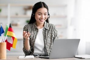 Happy asian lady student using laptop and headset, learning foreign language online, looking at notebook screen, talking and gesturing, taking notes while having chat with tutor, copy space photo