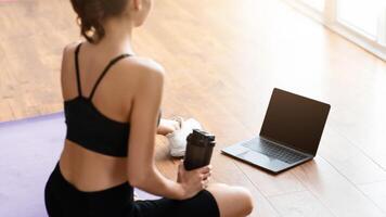 Rear back over the shoulder view of unrecognizable woman sitting on yoga mat drinking water and looking at blank laptop screen, having call with trainer or attending fitness class online, mockup photo