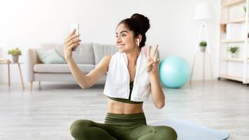 Joyful Indian woman taking selfie on break from home workout, full length. Happy Asian lady with smartphone making photo of herself on domestic training, making peace gesture and smiling at webcam