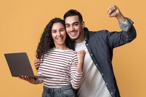 Online Win. Portrait Of Overjoyed Arabic Couple Celebrating Success With Laptop, Cheerful Middle Eastern Spouses Holding Computer, Raising Fists And Exclaiming With Excitement On Yellow Background photo