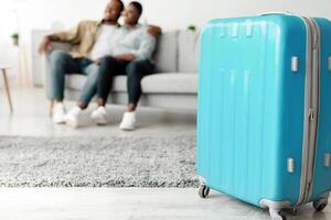 Blue Travel Suitcase Standing On Floor While Unrecognizable Black Couple Hugging Sitting On Couch Ready For Vacation Trip At Home. Tourists Waiting For Journey. Selective Focus On Luggage photo
