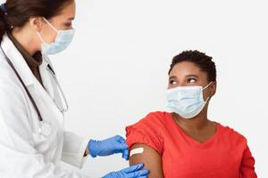 Covid-19 Vaccination. Black Woman Getting Vaccinated Against Coronavirus, Doctor Sticking Adhesive Plaster Strip After Corona Virus Vaccine Injection Shot On White Studio Background, Wearing Face Mask photo