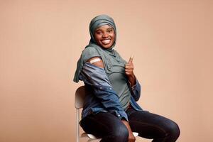 Antiviral Immunization, Vaccination. Black Muslim Lady In Hijab Vaccinated Against Covid-19 Showing Vaccinated Arm And Like Gesture Approving Quality Of Vaccine After Injection On Beige Background photo