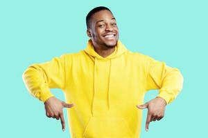 Look There. Happy Black Guy Pointing Fingers Down Standing On Blue Studio Background, Smiling To Camera. Bright Advertisement Banner With African Man Wearing Yellow Hoodie. Check This Offer photo
