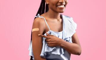 Cropped view of young black lady pointing at adhesive plaster om her arm after coronavirus immunization over pink studio background. Successful covid vaccination concept photo