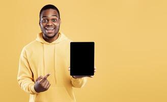 Black Man Pointing At Digital Tablet Empty Screen Holding Computer Posing Over Yellow Studio Background, Smiling To Camera. Mockup For Great Website Or Applicatioin Advertisement photo