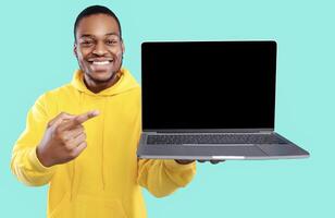 African American Guy Showing Laptop Computer Empty Screen To Camera Recommending Website Posing Over Blue Studio Background. Technology, People And Gadgets Concept. Mockup photo