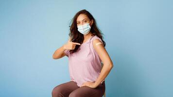 Antiviral Vaccination. Vaccinated Woman Wearing Face Mask Pointing At Arm Showing Plaster After Covid-19 Vaccine Injection Sitting On Blue Background. Coronavirus Immunization. Panorama, Free Space photo