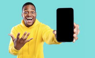 Wow Application. Excited African American Man Showing Phone With Blank Screen For Mobile App Advertisement Smiling To Camera In Excitement Over Blue Background. Studio Shot, Selective Focus, Mockup photo