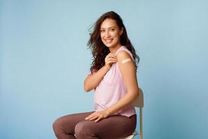 Coronavirus Vaccination. Happy Young Lady Showing Vaccinated Arm With Medical Adhesive Bandage Strip After Covid-19 Vaccine Injection Over Blue Studio Background. Covid Virus Immunization Campaign photo