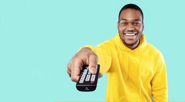 Cheerful Black Guy Watching TV Pointing Television Remote Controller To Camera Switching Channel Over Blue Studio Background. Happy Man Enjoying Movie. Selective Focus photo