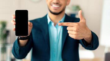 Cheerful young Arab businessman pointing at smartphone with blank screen at modern office, mockup for mobile app or website. Happy guy employee in formal wear showing blank cellphone with copy space photo