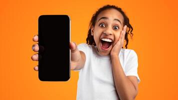 Wow, Amazing Offer. Portrait of excited African American girl holding mobile phone with empty screen in hand, showing device close to camera and touching cheek, mock up, yellow studio background photo
