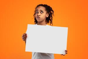 Portrait Of Pensive African American Girl In Doubt Holding And Showing Blank Square Paper Poster For Your Text Looking Up And Thinking, Standing Isolated Over Yellow Studio Background, Free Copy Space photo