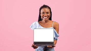 Funky black lady holding laptop pc with blank screen for your ad or website on pink studio background. Cool Afro woman showing portable computer with mockup for design or advertisement photo