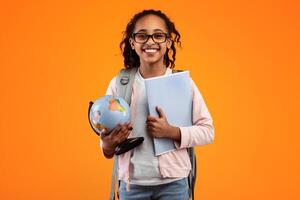Portrait of cheerful African American girl in glasses holding Earth world globe and notebook, wearing backpack and looking at camera isolated on yellow studio background. Education in school concept photo