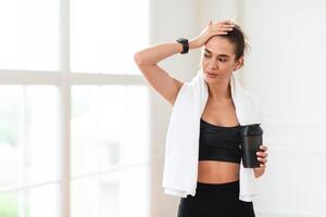 After Training. Tired young woman with white towel on neck holding shaker with water swiping sweat from forehead resting after training routine standing indoors at gym or home photo